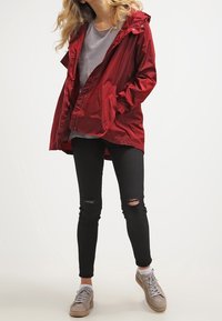 Czerwona wodoodporna kurtka z kapturem, z zamkiem z przodu i bocznymi kieszeniami, noszona na szarym T-shircie. Czarne przetarte dżinsy skinny i jasnoszare trampki.