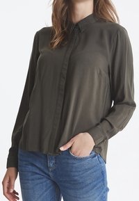 Chemise olive verte à boutons, fabriquée en tissu lisse, avec des manches longues et un col. Ajustement légèrement décontracté avec une poche poitrine.