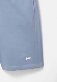 Shorts bleu clair en tissu doux, avec des poches latérales, une texture lisse et un logo blanc en bas à gauche de l'ourlet.