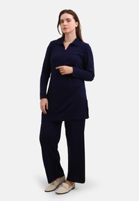 Navy blue, long-sleeve tunic with a collared neckline and side slits, paired with matching wide-leg pants and light-colored loafers.