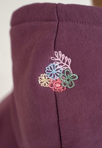Lila szövet texturált felülettel, kék, rózsaszín, zöld és sárga hímzett virágokkal a nyakkivágásnál.