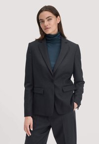 Zwarte op maat gemaakte blazer met enkele knoopsluiting, notch revers, twee voorkruiken, gecombineerd met een donkere coltrui. Glad materiaal en gestructureerde pasvorm.