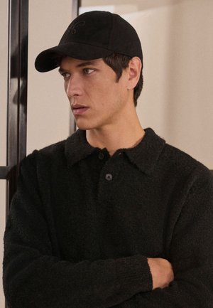 Giovane uomo che indossa un pullover nero strutturato e un cappellino da baseball nero, in piedi con le braccia incrociate, guarda verso sinistra al chiuso.