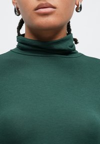 Πράσινο ζιβάγκο από απαλό, ελαστικό ύφασμα, με ψηλό γιακά και ριγωτή υφή, συνοδευόμενο από ασημένιο σκουλαρίκι-κρίκο.