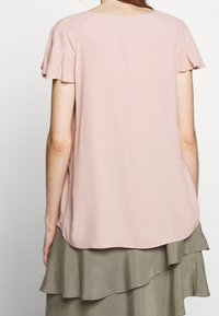 Różowa, krótko rękawowa bluzka z falbanami; miękki, lekki materiał i zaokrąglony dół. Noszona z warstwową, oliwkowozieloną spódnicą.