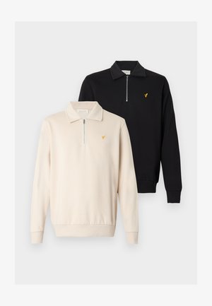 Zwei Sweatshirts mit Viertel-Reißverschluss: eines in Creme mit Fronttasche und gelbem Logo, das andere in Schwarz mit gerippten Bündchen und Kragen.