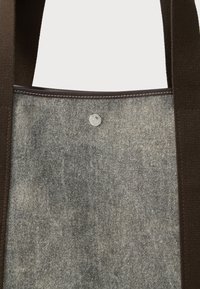 Τσάντα tote από γκρι υφασμάτινη επιφάνεια, με σκούρους καφέ ιμάντες και κλείσιμο με ασημί κουμπί snap κοντά στο κέντρο της κορυφής.