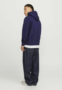 Felpa con cappuccio blu navy dal fit rilassato, abbinata a pantaloni di denim scuro e sneaker chiare. Il tessuto sembra morbido, con un design minimalista.