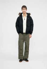 Μαύρο μπουφάν με μόνωση και κουκούλα επενδεδυμένη με γούνα, μπροστινό φερμουάρ και δύο μεγάλες τσέπες, συνδυασμένο με ελαιόχρωμες cargo παντελόνες και μαύρα αθλητικά παπούτσια.