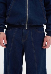 Zestaw ciemnoniebieskiej kurtki i spodni dżinsowych z zamkiem błyskawicznym, prążkowanymi mankietami oraz kontrastowym pomarańczowym stębnowaniem, z kieszeniami bocznymi.