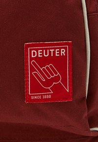 Étiquette en tissu rouge avec le texte blanc "DEUTER" et un symbole de main. Bordure cousue avec "SINCE 1898" en dessous du logo, fixée à un matériau rouge plus foncé.