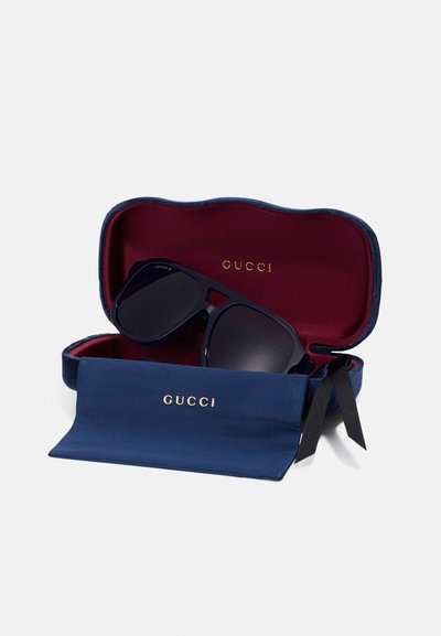 Étui à lunettes de soleil navy avec un extérieur texturé, doublure intérieure rouge et logo "GUCCI" doré. Les lunettes de soleil à l'intérieur présentent des verres foncés et un style de monture audacieux.