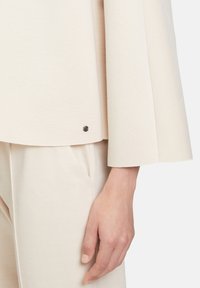 Giacca in misto lana color crema con maniche larghe, dotata di dettaglio a pressione argentato all'orlo; abbinata a pantaloni leggeri a spina di pesce.