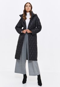 Długie czarne pikowane płaszcz z kapturem, z diamentowym wzorem, dwoma przednimi kieszeniami i eleganckim fasonem, zestawione z szerokimi szarymi spodniami.