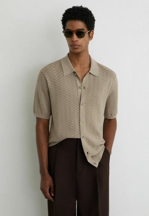 Mees, kes kannab mustrilist beeži lühikeste varrukatega nööpidega särki, tumepruune pükse ja päikeseprille, seisab valge seina ääres.