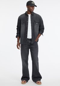 Cienka, dopasowana koszula z ciemnego dżinsu nałożona na białą koszulkę, w połączeniu z ciemnymi dżinsami i kolorowymi trampkami. Akcesoria to ciemna czapka.