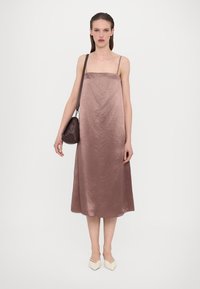 Satin midi-kjole i mauve med tynne stropper, en avslappet passform og en glatt tekstur. Modellen bærer en liten mørkebrun håndveske og har på seg kremfargede spisse sko.