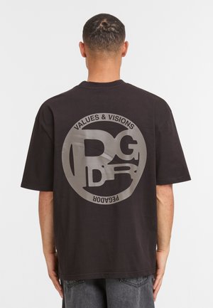 Man gezien van achteren, gekleed in een zwart T-shirt met een groot cirkelvormig grijs logo en de woorden "VALUES & VISIONS PEGADOR" erop.