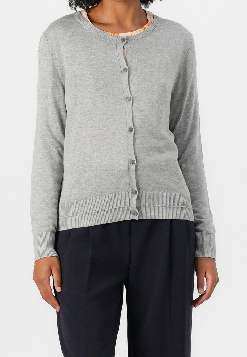 Femme portant un cardigan gris clair boutonné sur un haut fleuri et un pantalon noir, debout devant un fond uni.