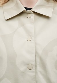 Κοντινό πλάνο σε μπεζ σακάκι με διακριτικό μεγάλο κυκλικό σχέδιο, κλείσιμο με κουμπιά και μυτερό γιακά πάνω από μαύρο τοπ.