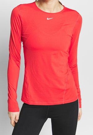 Femme portant un haut de sport Nike rouge vif à manches longues et un legging noir, debout avec une main le long du corps.