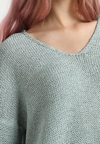 Svetle zelený pletený svetr s výstřihem do V, volným střihem a texturovaným vzorem, vyrobený z měkké, prodyšné látky.