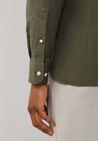 Groen shirt met lange mouwen en een manchetknoop. Heeft een gladde textuur, afgeronde zoom en witte knoopdetails op de mouwen.