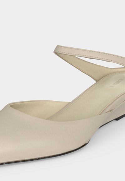 Ballerine beige à bout pointu avec fine bride cheville et texture cuir lisse, inclinée pour montrer les détails du côté et de la semelle intérieure.