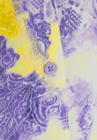 Tessuto a fantasia con tonalità di viola e giallo, con un motivo paisley e un dettaglio di bottone lilla. Materiale in cotone testurizzato.