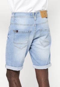 Short en jean bleu clair avec revers roulés porté par une personne, montrant les poches arrière et un patch de marque en cuir marron sur la ceinture.