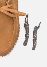 Zapato de cuero marrón claro con una superficie texturizada y suela de goma, acompañado de dos cordones grises con un patrón espiral blanco.