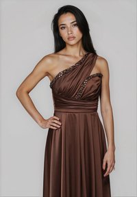 Donna con lunghi capelli scuri che indossa un abito in satin marrone con una spalla e rifiniture di paillettes, in piedi con una mano sulla vita su uno sfondo neutro.