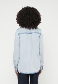 Light blue denim shirt with a button closure, long sleeves, rounded hem, and contrasting stitching on shoulders and cuffs.