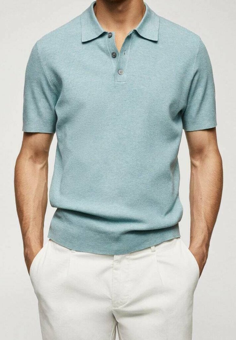 Homme portant un polo en maille bleu clair à manches courtes et un pantalon blanc, les mains dans les poches, sur un fond uni.