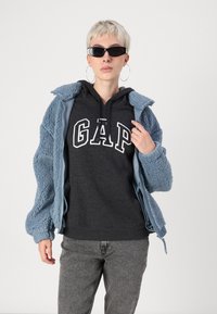 Jachetă din fleece pufoasă, de culoare albastru deschis, cu fermoar, purtată peste un hanorac gri închis cu inscripția albă "GAP". Ochelari de soare negru în formă de ochi de pisică. Blugi gri.
