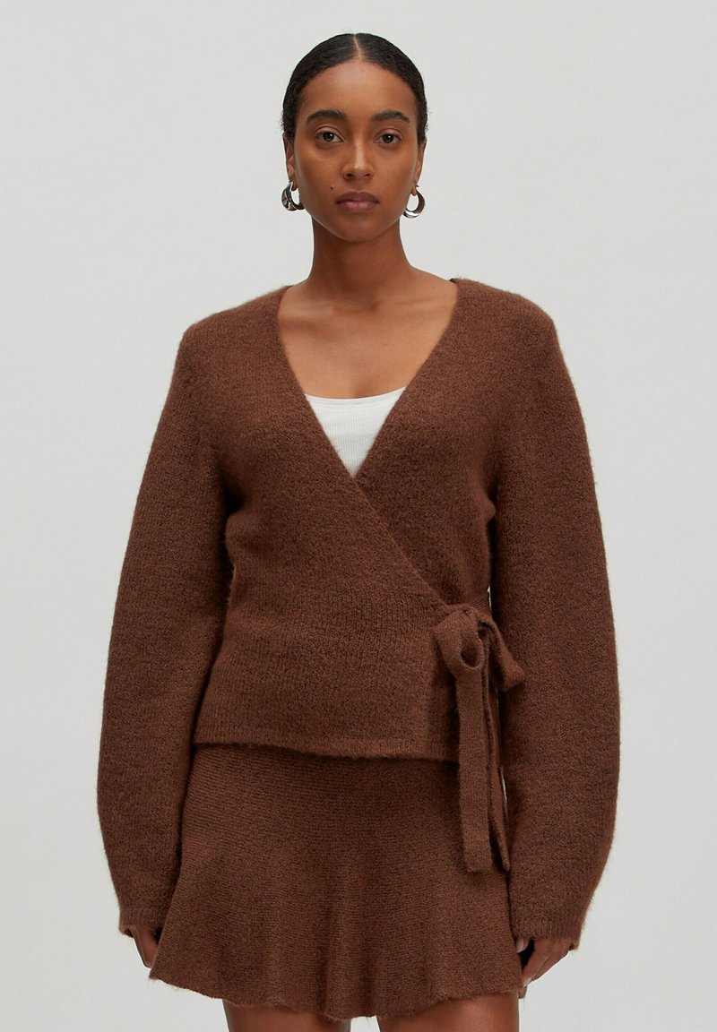 Bruine, fuzzy wikkelcardigan met lange mouwen en een strikdetail in de taille, gecombineerd met een bijpassende minirok. Zachte textuur en diepe V-hals.