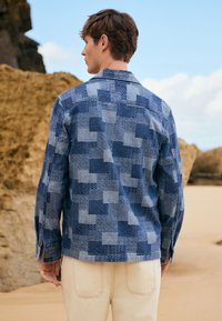 Giovane uomo in piedi all'aperto di spalle, indossa una giacca blu con motivo e pantaloni beige chiaro, con grandi formazioni rocciose sullo sfondo.