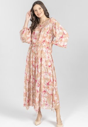 Kobieta uśmiechająca się, ubrana w długą beżową, zwiewną sukienkę w różowo-białe kwiatowe wzory, stojąca z jedną ręką na biodrze, a drugą przy włosach.