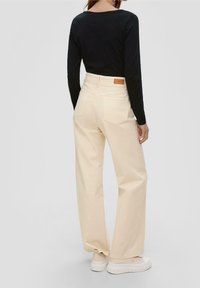 Beigefarbene, weit geschnittene Hosen aus Baumwolle mit Gesäßtaschen und einem Lederschild am Bund. Kombiniert mit weißen Sneakers und einem schwarzen, langärmligen Oberteil.