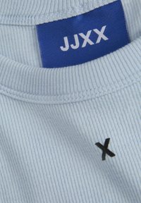 Světle modré žebrované tričko s modrou etiketou, na které je vytištěno "JJXX" a malá černá výšivka 'X' na látce. Hladká textura.