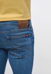 Siniset farkut, joissa on istuva malli. Farkuissa on ruskea nahkapaikka, jossa lukee "MUSTANG", sekä taskuissa erottuvat punaiset ja keltaiset tikkaukset.