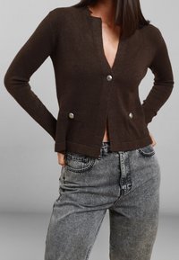 Cardigan marrone realizzato in materiale morbido, con scollo a V, bottoni argento e tasche laterali. Indossato sopra jeans grigi a vita alta.