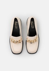 Mocassins beiges à finition en cuir lisse, ornés d'un accent en chaîne dorée sur le dessus et d'une semelle plate en caoutchouc noir.