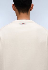 Sudadera beige con cuello redondo y un pequeño detalle bordado con rayas rojas, blancas y azules en la parte trasera del cuello.