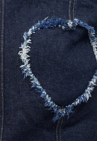 Σκούρο μπλε τζιν ύφασμα με ξεφτισμένο, καρδιόσχημο μπαλώμα που δείχνει πιο ανοιχτά εσωτερικά νήματα και κίτρινα διακοσμητικά ραψίματα.