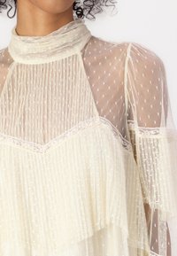 Sheer cream blouse with textured polka dots, ruffled hem, and lace accents at the neckline and sleeves. Soft and lightweight fabric.