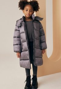 Geacă puffer gri cu glugă, având cusături orizontale și un fermoar frontal. Purtați-o peste un top închis la culoare și asortați-o cu cizme negre cu șireturi.
