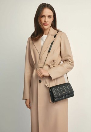 Kobieta o brązowych włosach do ramion nosi beżowy trencz z paskiem i niesie czarną pikowaną skórzaną torbę na ramię z złotym łańcuszkiem.