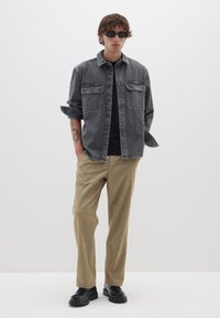 Camicia in denim grigia con due tasche sul petto, t-shirt nera sotto, pantaloni beige e scarpe nere. Modello che indossa occhiali da sole neri.