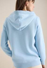 Femme de profil portant un sweat à capuche bleu clair à manches longues, sur un fond beige uni.